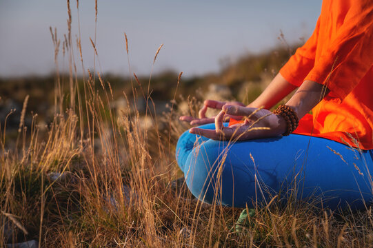 Close-up of a woman and female hands sitting in a lotus position, yoga outdoors in the grass at sunset against the backdrop of a mountain range and clouds, meditation