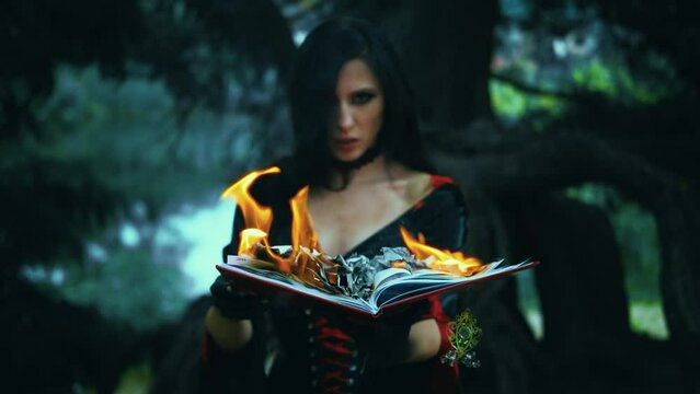 Fantasy woman sexy witch holds old burning book in hands, bright flame fire, Girl fairytale goth reads magic spell. Black red dress halloween gothic costume. Dark deep forest tree. Light on evil face