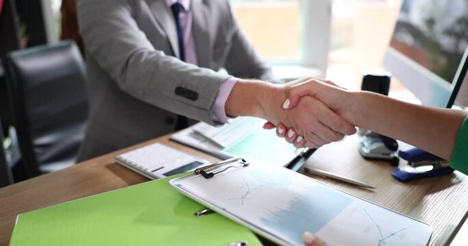Two business people shake hands sitting at table with financial report charts