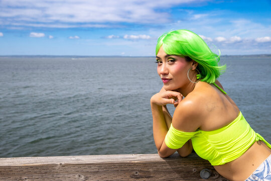 Woman with vibrant neon green hair and matching top leans on the rail of an ocean front dock. There is an expansive view of the water on a pretty summer day. 