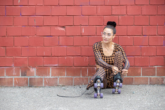 Asian woman sitting on the ground against a red brick wall. She is tying the laces on her roller skates while wearing a checkered body suit. 