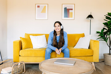 Happy young millennial woman sitting on sofa while listening to music with headphones