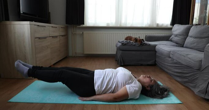 Senior woman doing pilates exercises on yoga mat, lifting and strengthening waist, her dachshund dog in the background. Concept of exercises for mature people at home and healthy lifestyle