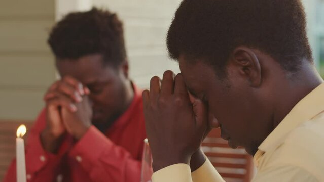 Waist up of two young Black men sitting with their heads down at dinner table in summerhouse praying