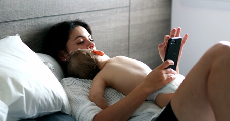 Casual mother looking at cellphone with baby napping in bedroom, infant asleep on mom chest