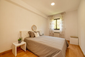 bedroom with double bed with headboard with gray cushions and round mirror with wooden frame and brown aluminum window with floral curtains