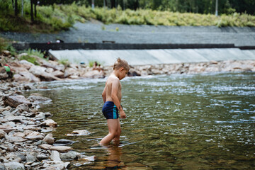 A child enters the water, summer holidays on the river, a boy alone bathes in the cold water of a mountain river, a stone bed, a summer vacation in nature, swimming trunks on the body