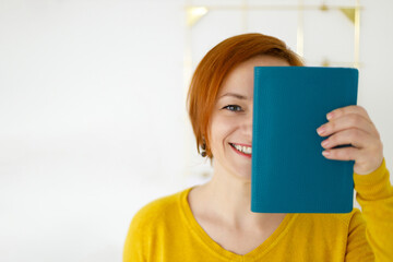 woman covered her face with diary. Business red haired woman in yellow blouse holding notebook in her hand and smiling. Business planning and tasks for the day or week. Female in business