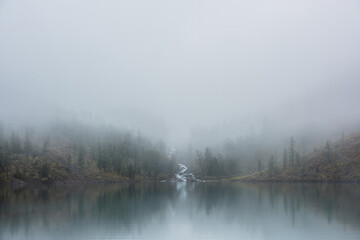 Mountain creek flows from forest hills into glacial lake in mysterious fog. Small river and coniferous trees reflected in calm alpine lake in misty morning. Tranquil landscape in fading autumn colors.