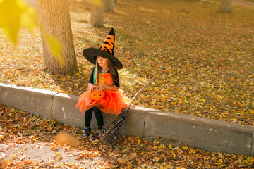 A cute girl dressed up in a witch costume is sitting on the outskirts of the sidewalk, covered with...