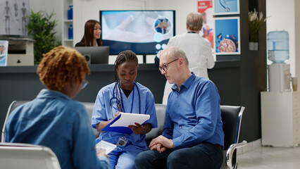 African american nurse consulting retired patient in waiting area, taking notes on report papers to...