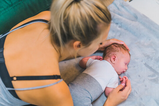 Adorable newborn baby boy with cute brown hair, sleeping peacefully on his stomach on a bed at home. Young caucasian mom gently touching his head and hugging her son. High quality photo