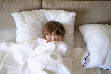 Happy little boy talking on smartphone, looking aside while sitting on bed