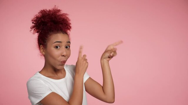 Close up cheerful young African American redheaded curly woman pointing finger to side workspace promo area mock up and presenting product. Portrait happy female standing on isolated pink background