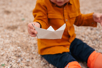 toddler boy playing with paper boat wearing rubber boots on the beach near sea in autumn or summer time
