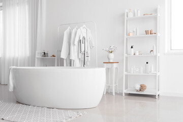 Interior of stylish bathroom with modern bathtub and shelf units with accessories near white wall