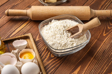 Glass bowl with flour and scoop on wooden background