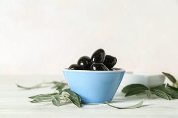 Bowl of tasty black olives and leaves on light wooden table