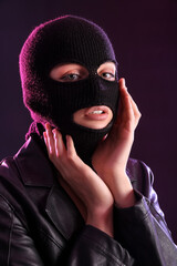 Young woman in balaclava and leather jacket with hands near face against dark background