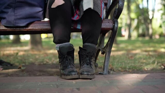 Legs of unrecognizable homeless woman in dirty clothes and boots pouring off leftovers of coffee from cup. Desperate Caucasian young beggar sitting on bench in sunny summer park