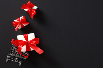 Shopping cart and white gift boxes with red ribbon bows on black background. Black Friday Sale banner design. Top view with copy space.