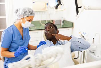 Upset african american man sitting in dental chair, complaining to qualified female dentist about toothache.. - obrazy, fototapety, plakaty