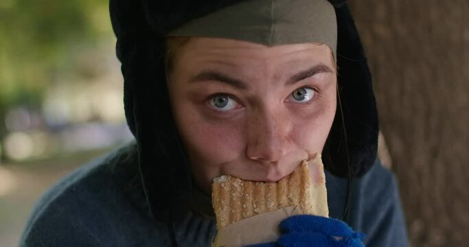 Close-up hungry homeless young woman eating bun in slow motion looking at camera with grey eyes. Headshot portrait of starving sad Caucasian poor beggar posing in summer park. Cinema 4k ProRes HQ