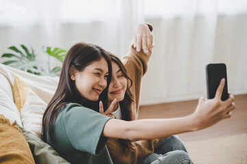 Two asian young women happy smiling and taking selfie on couch in living room at home