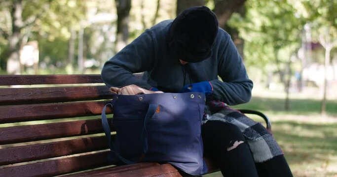 Focused homeless young woman rummaging in bag in slow motion sitting on bench in sunny park. Portrait of sad Caucasian poor houseless person searching food outdoors. Cinema 4k ProRes HQ