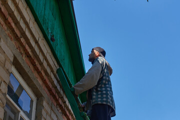A man in a baseball cap stands on the wooden stairs of a village house. There is a padlock on the attic door. The man looks thoughtfully at the door.