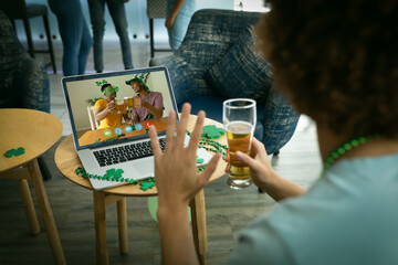 Mixed race man holding beer at bar making st patrick's day video call waving to friends on laptop