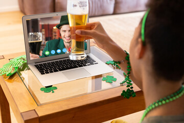 Mixed race woman holding beer having st patrick's day video call with male friend on laptop at home