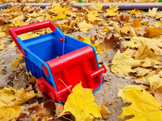 Red and blue plastic toy scoop in the sand on a playground with yellow leaves