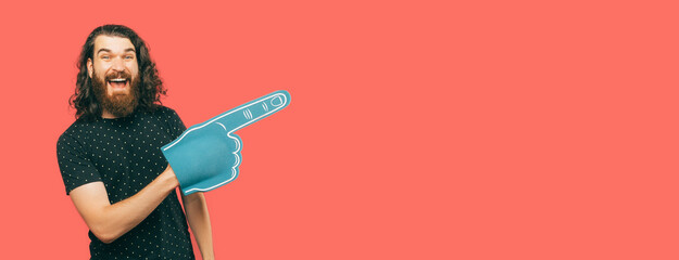 Excited bearded man is pointing aside while wearing blue fan foam glove. Studio shot over coral background with copy space.