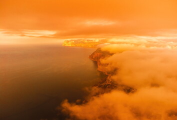 A red burning sunset over the sea with rocky volcanic cliff. Abstract nature summer or spring ocean sea background. Small waves on golden warm water surface with bokeh lights from sun.