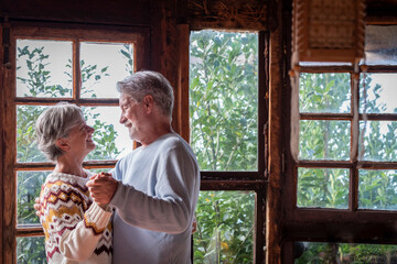 Old man and woman happy couple dancing at home in living room with outdoors background and windows....