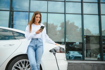 Happy young adult woman smiling wide, looking away, charging automobile battery from small public station, standing near electric car.