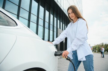 Smiling young caucasian woman plugging electricity cable in electric vehicle for charging on sunny...