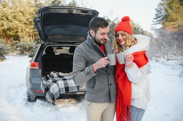 Couple in love sitting in car trunk drinking hot tea in snowy winter forest and chatting. People relaxing outdoors during road trip. Valentines day