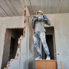 Full length of man in workwear drilling wall with hammer drill. Male worker using drill breaker...