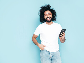 Handsome smiling hipster  model.Sexy Arabian man in summer stylish clothes.Fashion male with long curly hairstyle posing near blue wall in studio.Looking at cellphone screen. Using apps