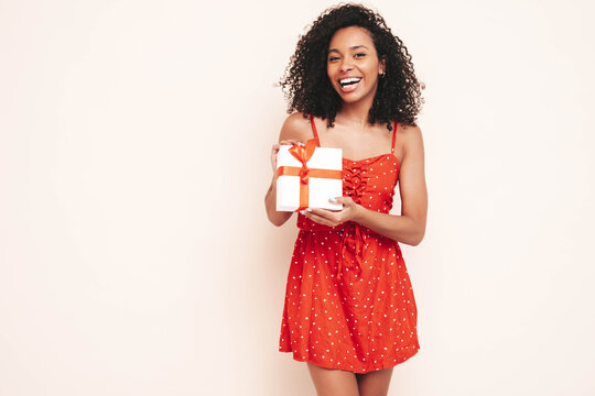 Beautiful black woman with curls hairstyle. Smiling model dressed in summer red dress. female posing near wall in studio. Tanned and cheerful. Holding gift box. Shocked, surprised