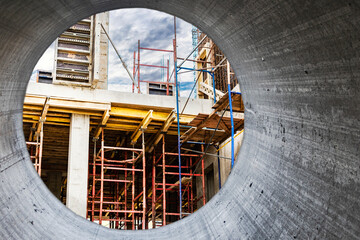 Obraz premium Construction of a new monolithic reinforced concrete house. Scaffolding on the facade of a building under construction. Working at height on a construction site. View from a large concrete pipe.