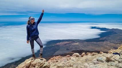 Front view on happy woman standing on rock enjoying panorama from summit of volcano Pico del Teide on the island of Tenerife, Canary Islands, Spain, Europe. Island covered in cloud. Freedom concept