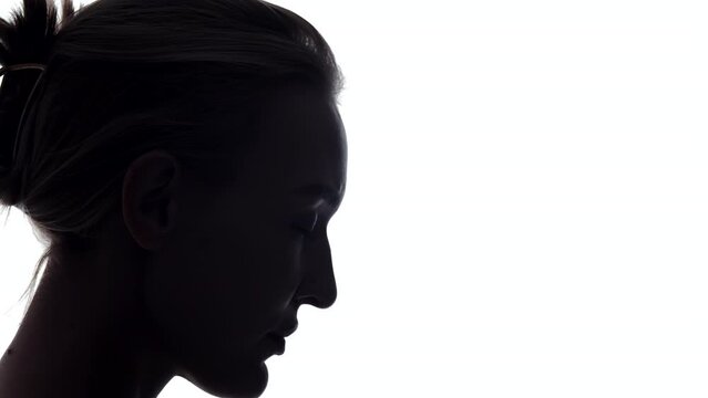 Side view closeup portrait of an attractive young caucasian woman with her eyes closed, slowly lowering her head down. Silhouette, in the shadow, white background. Meditation, mindfulness concept