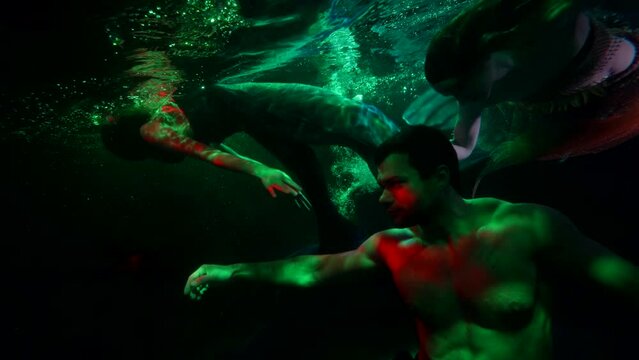 a man with a naked torso is floating in dark water. a woman and a mermaid swim in the background. green and red light
