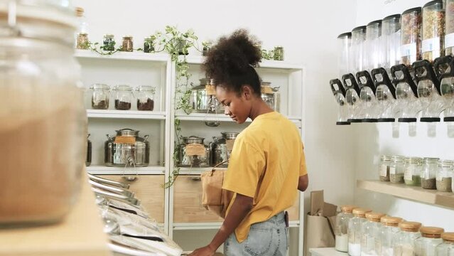 Young African American woman is choosing and shopping for organic products in refill store with reusable bag, zero-waste grocery, and plastic-free, eco environment-friendly, sustainable lifestyles.