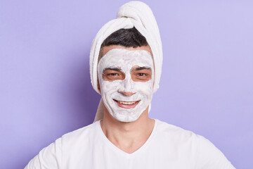 Portrait of smiling Caucasian man with mask for skin, standing wrapped in towel isolated over violet background, doing cosmetology procedures at home.