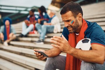 Disappointed sports fan watching soccer game on mobile phone outdoors.