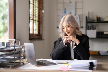 Obraz premium Young finance market analyst working at sunny office on laptop while sitting at wooden table. Businesswoman analyze document graphs and diagramm on notebook screen
